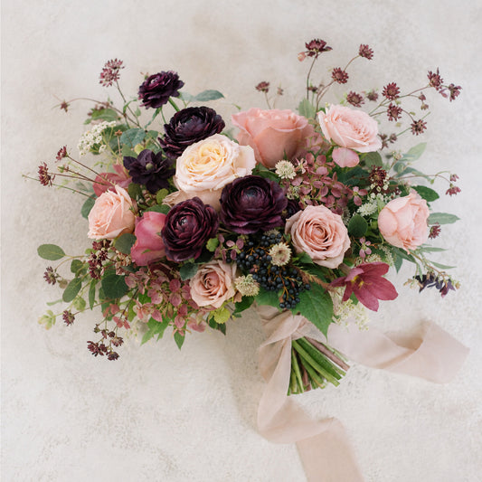 A flatlay of moody bridal bouquet with burgundy, blush, mauve, and antique peach garden flowers wrapped in a chiffon ribbon