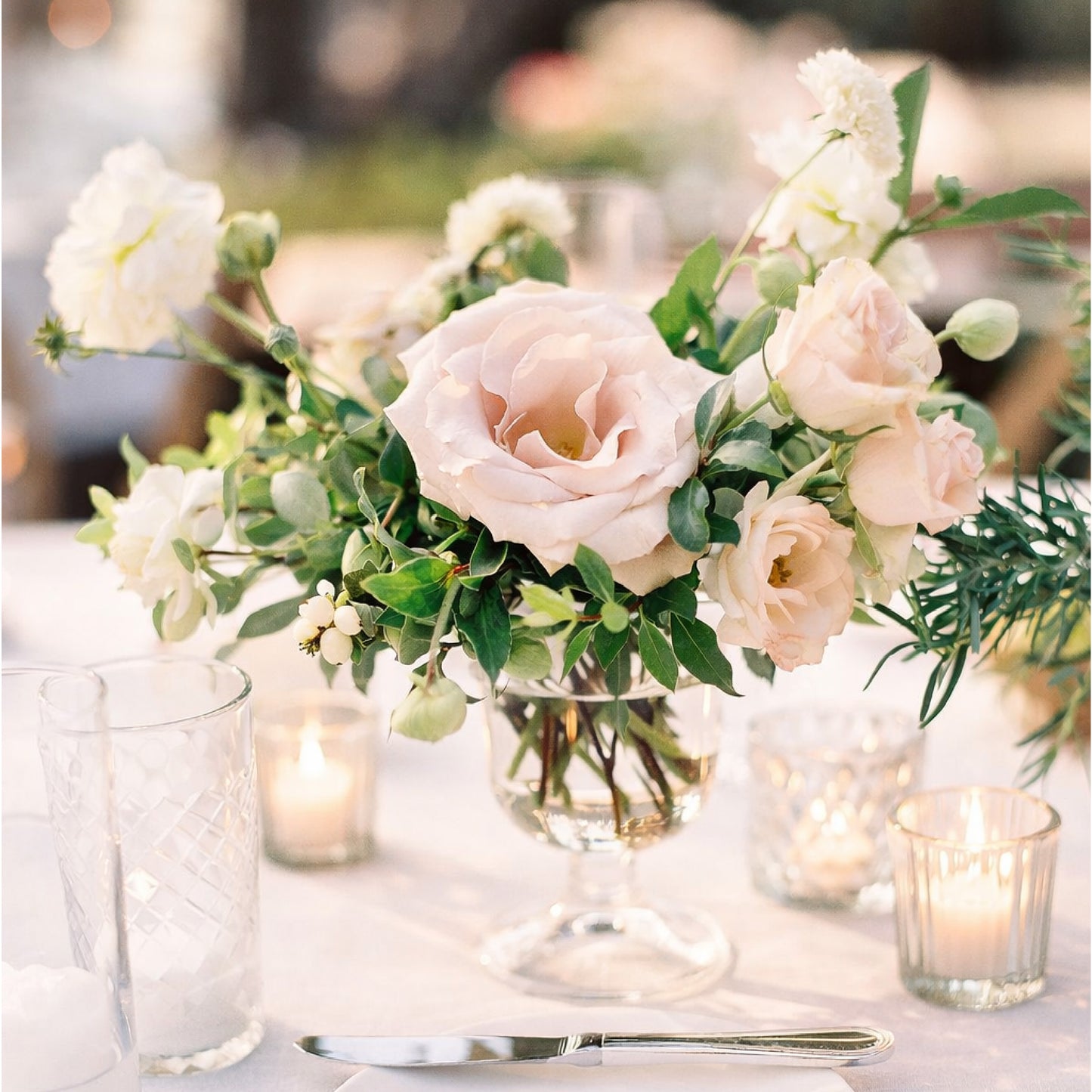 Soft blush and ivory floral arrangement in a glass compote vase styled on a romantic candlelit reception table