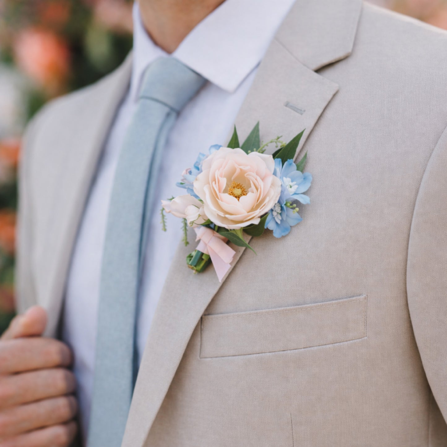 Pastel wedding boutonnière with blush rose and powder blue flowers on beige suit.