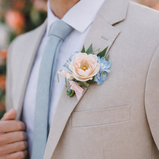 Pastel wedding boutonnière with blush rose and powder blue flowers on beige suit.