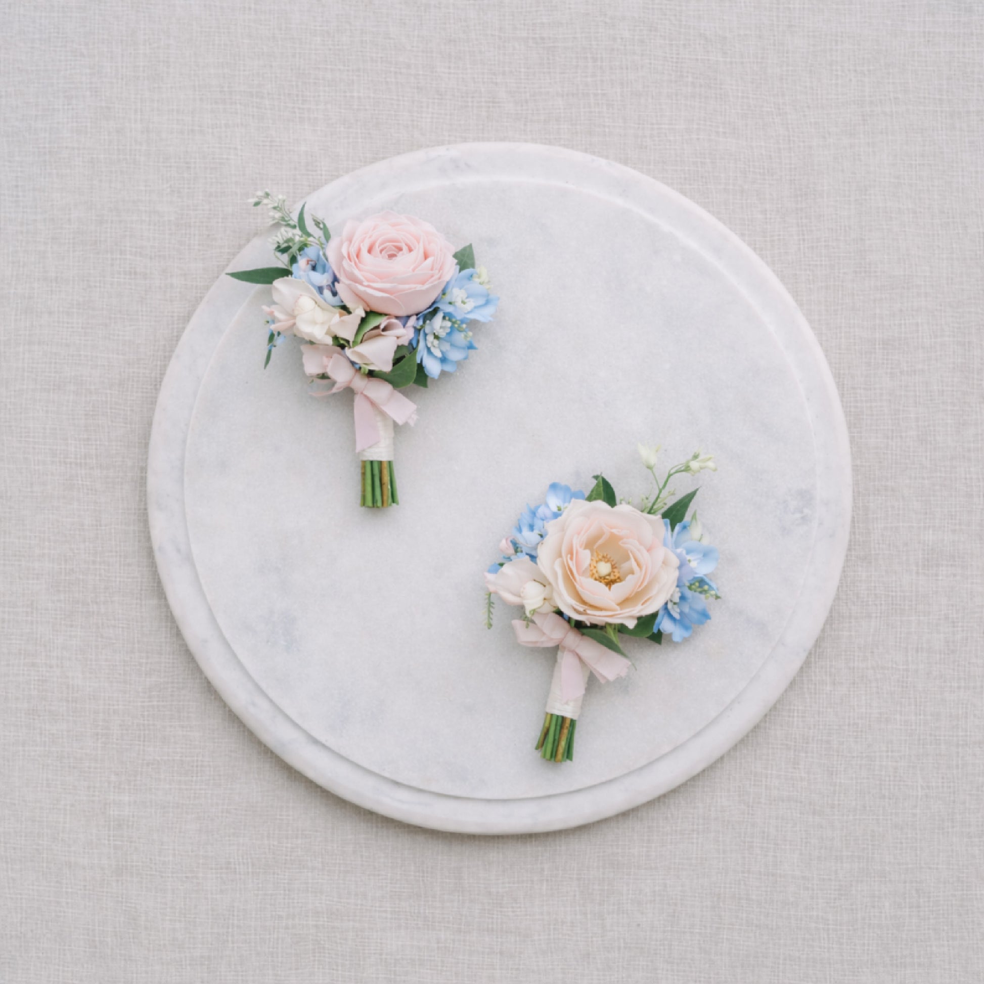 Close-up of pastel wedding boutonnières with blush roses and powder blue flowers displayed on marble.