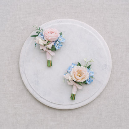 Close-up of pastel wedding boutonnières with blush roses and powder blue flowers displayed on marble.