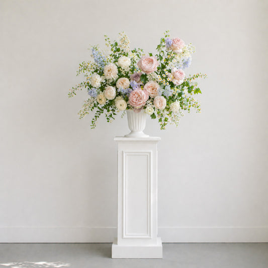 Pastel ceremony urn arrangement displayed on pedestal with blush, blue, lavender, and ivory garden flowers