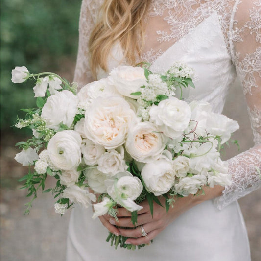 Porcelain bridal bouquet with white garden roses, ranunculus, and airy seasonal blooms held by bride in lace wedding gown.