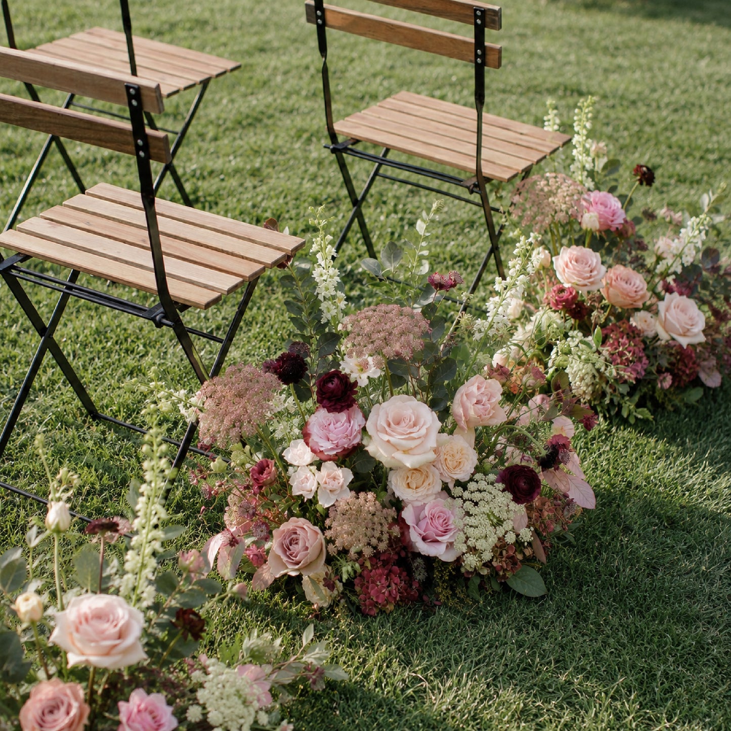 Moody burgundy and blush meadow arrangements styled along outdoor wedding aisle