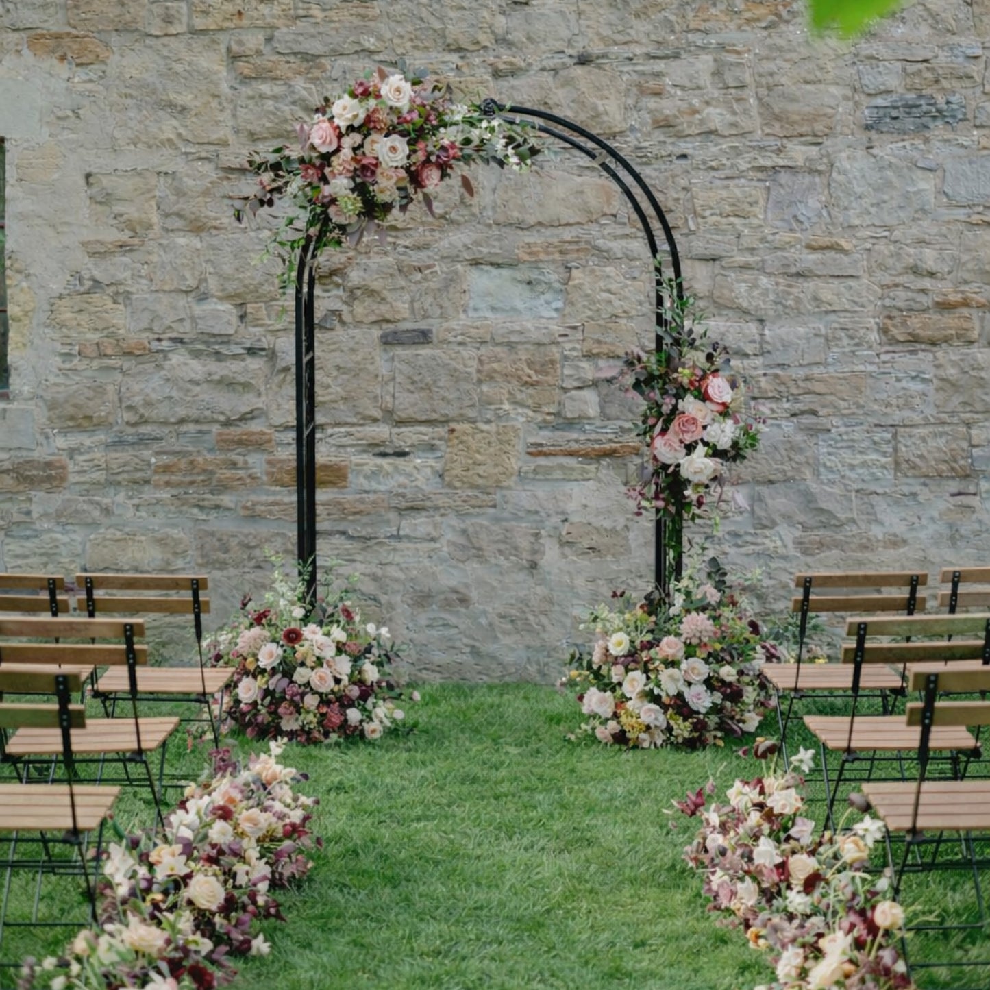 Moody burgundy and blush floral swags framing outdoor wedding ceremony arch with ground Meadow arrangements at base of arch and along the aisle