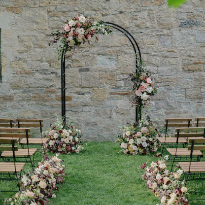 Moody burgundy and blush floral swags framing outdoor wedding ceremony arch with ground Meadow arrangements at base of arch and along the aisle