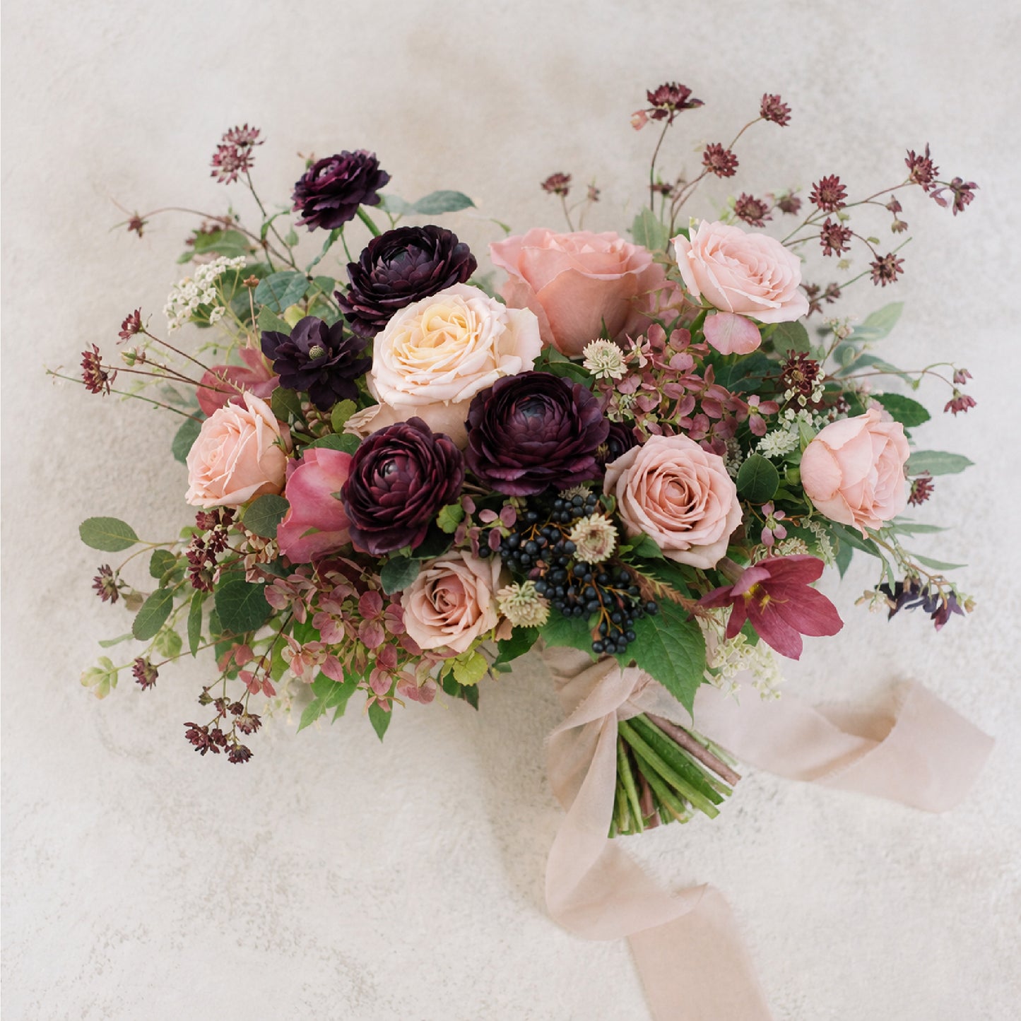 A flatlay of moody bridal bouquet with burgundy, blush, mauve, and antique peach garden flowers wrapped in a chiffon ribbon