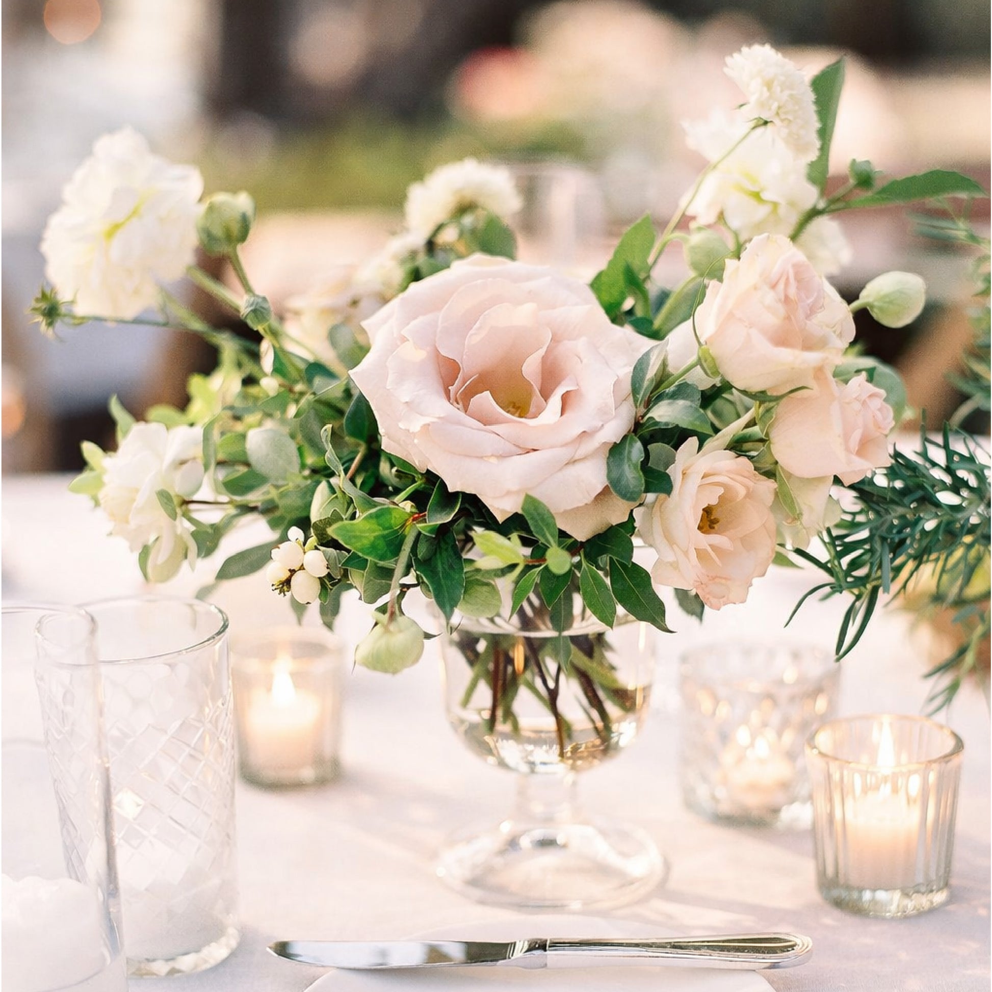 Soft blush and ivory floral arrangement in a glass compote vase styled on a romantic candlelit reception table